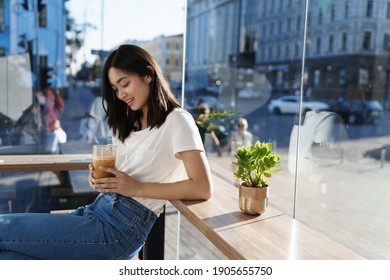 Vedere laterală a unei femei asiatice fericite stând singură în cafenea lângă fereastră, zâmbind în timp ce bea latte cu gheață în ziua însorită de vară, purtând tricou cu blugi.
