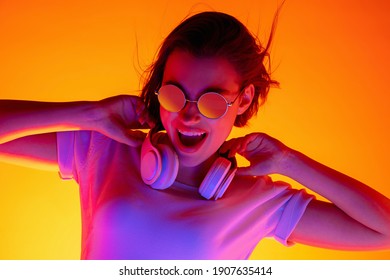 Conducir. Retrato de mujer caucásica sobre fondo de estudio naranja en luz de neón rojo-rosa. Hermosa modelo femenina con gafas de sol y dispositivo. Concepto de emociones humanas, expresión facial, ventas, publicidad, moda.