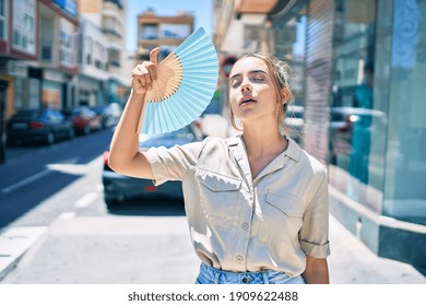Jovem linda mulher loira caucasiana sorrindo feliz ao ar livre em um dia ensolarado usando leque para o tempo quente