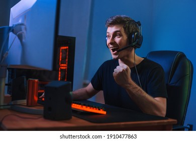 若い白人のプロゲーマーがオンラインビデオゲームで勝利し、幸せを感じて終了し、はいの手のジェスチャーを示します。自宅でヘッドフォンを使用してコンピュータでトーナメントを行うプロのサイバースポーツマン、赤と青