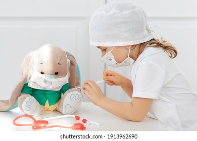 小さな子供の女の子は、おもちゃに注射を行います。医者を演じる医療用マスクを持つかわいい子、ワクチンの入った注射器を持っています。コロナウイルスの予防接種のコンセプト。子供は自宅でロール ゲームをプレイします。