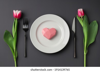Kreative Tischdekoration mit Herzen auf einem weißen Teller, schwarzer Gabel und Messer, Tulpen auf dunklem Hintergrund. Valentinstag, Hochzeitstag, Geburtstag, Frauentag und Muttertag. Flach liegen