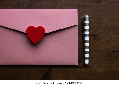 A salmon coloured envelope holding a wedding invitation with a red felt heart as the seal, next to a small gold and white pen on a dark wooden desk, Toronto, Ontario, Canada.