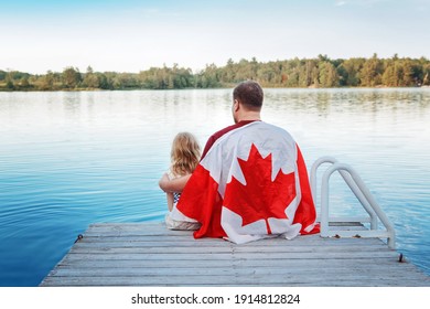 父と娘は、湖のそばの木製の桟橋に座って大きなカナダの旗に包まれました。カナダの日のお祝い屋外。7 月 1 日、カナダの日を祝って一緒に座っているお父さんと子供。