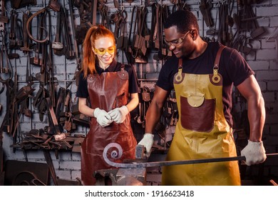 couple in love redhaired ginger young european woman and afro-american man wearing leather apron working blacksmith workshop.small family international business concept