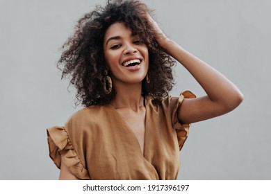 Retrato de una mujer africana morena rizada de piel oscura en la parte superior beige sonriendo y erizando el cabello sobre un fondo gris aislado.