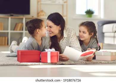Nette Familie, die den Muttertag zu Hause feiert. Glückliche junge Frau, die mit ihren Kindern auf dem Boden liegt, Grußkarte liest, Kindern für das Geschenk dankt, Nasen berührt und Eskimo mit ihren Töchtern küsst
