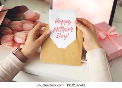 Die Hände der Frau, die einen Brief im Handwerksumschlag halten. Rosa Hintergrund, Muttertagskonzept. Tulpen blühen und rosa Geschenkbox im Hintergrund. Schreibtisch für damen zu hause.