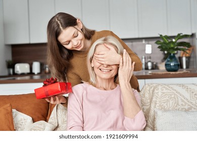 Una linda niña adolescente, una hija pequeña que se divierte cubriendo los ojos de mamá sosteniendo una caja de regalo roja que hace que el Día de la Madre sea una sorpresa para saludar a la feliz mamá o abuela en las vacaciones de primavera en casa.