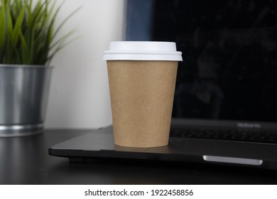 A take-away paper cup rests on a black wooden table, a computer and an office plant. This photo is suitable for use as a mockup to put your logo or design