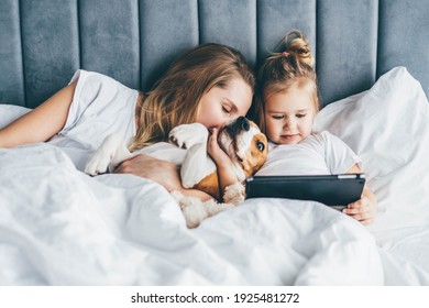 女の赤ちゃんと母親と犬がベッドに横たわり、タブレットで見ている。一緒に時間を過ごす家族。