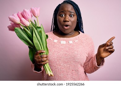Junge afrikanisch-amerikanische Plus-Size-Frau mit Zöpfen, die einen Blumenstrauß aus rosa Tulpen halten Überrascht, mit dem Finger zur Seite zeigend, offener Mund, erstaunter Ausdruck.