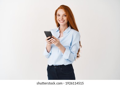 Happy pretty lady with ginger hair and blue eyes using mobile phone, smiling at camera, standing against white background.