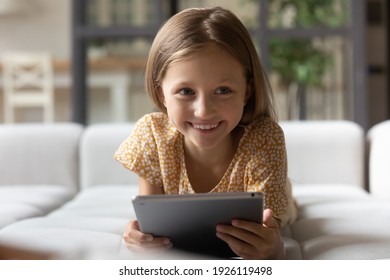 It is so cute. Laughing little girl lie on sofa having fun distracted from electronic tablet screen enjoy comic cartoon video. Cheerful kid studying easy with pleasure using funny education app on pad