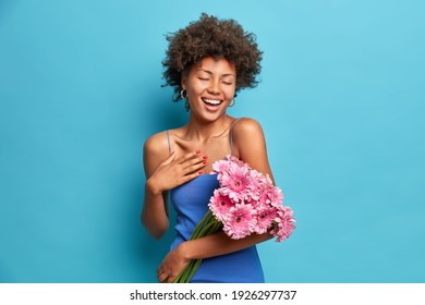 Das Foto einer erfreuten, aufrichtigen, dunkelhäutigen Frau, die angenehm lächelt, hält einen schönen Blumenstrauß aus Gerbera-Blumen und trägt ein Kleid, das über blauem Hintergrund isoliert ist. Frühlingsfrau hat festliche Stimmung. 8. März Konzept