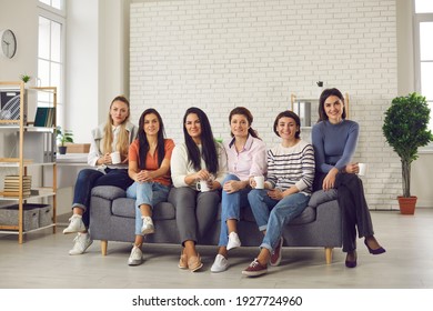 Happy friends sitting on sofa at home, enjoying nice get-together and cup of good coffee. Group of young women having break on Casual Friday at work, sitting on modern office couch and drinking tea