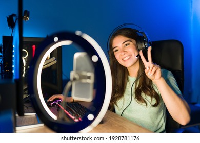 Portret van een mooie jonge vrouw die haar online kijkers gedag zegt tijdens een livestream met een smartphone en een ringlicht. Glimlachende vrouwelijke gamer die een videogame speelt op een gaming-pc