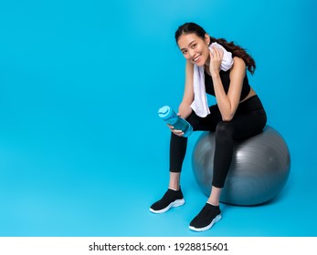 水のボトルを保持し、青い色の背景に分離された運動の後フィット ボールの上に座ってアジアの美しい幸せな女。スリムで健康的な女の子のトレーニングの概念。