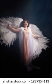 Chica morena con un vestido elegante y con alas de ángel blancas sobre un fondo negro. Modelo, actriz o bailarina posando en el estudio.