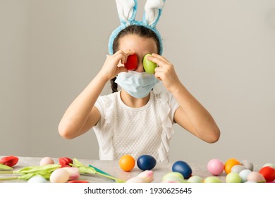 Coronavirus y Semana Santa. Niña linda en una máscara médica protectora con orejas de conejo con huevos de Pascua y flor de primavera.