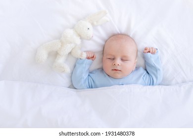 Bé trai ngủ trên giường nằm ngửa với đồ chơi nhồi bông thỏ trong bộ đồ ngủ màu xanh giơ tay, giấc ngủ trẻ sơ sinh khỏe mạnh
