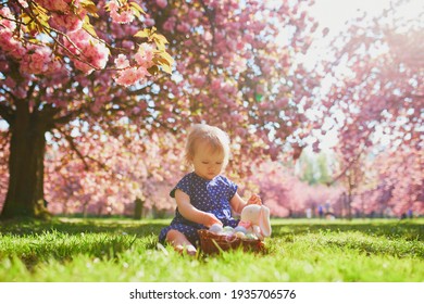Cute little one year old girl playing egg hunt on Easter. Toddler sitting on the grass under cherry blossom tree in full bloom. Little kid celebrating Easter outdoors in park or forest
