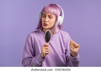 Young pretty woman with violet hairstyle singing and dancing with hair brush or comb instead microphone at very peri wall. Girl in headphones having fun, listening to music, dreams of being celebrity.