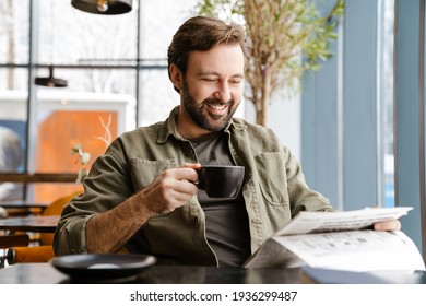 Unshaven smiling man drinking coffee while reading newspaper in cafe