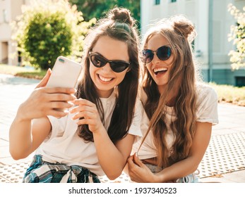 トレンディな夏の白い t シャツの服を着た 2 人の若い美しい笑顔の流行に敏感な女性の肖像画。通りの背景にポーズをとってセクシーな屈託のない女性。楽しんで、抱き合って、自撮りするポジティブなモデル