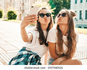 トレンディな夏の白い t シャツの服を着た 2 人の若い美しい笑顔の流行に敏感な女性の肖像画。通りの背景にポーズをとってセクシーな屈託のない女性。楽しんで、抱き合って、自撮りするポジティブなモデル