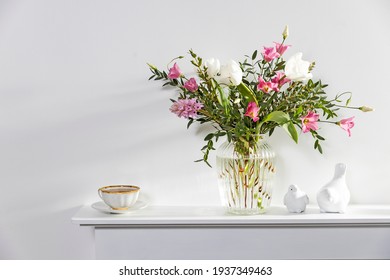 Een minimalistisch boeket van witte tulpen, roze eustoma, hyacint, eucalyptus in een gecanneleerde glazen vaas op een wit paneel van een kunstmatige open haard. Kopje thee, faience beeldjes van een twee vogels