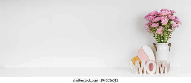 Moederdagdecor met een vaas met mamabloemen, houten MOM-letters en hart op een witte plank tegen een witte muurbannerachtergrond. Ruimte kopiëren.