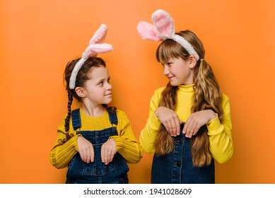 素敵なかわいい女の子はお互いにフレンドリーに見え、ウサギのように手を握り、ピンクのバニーのふわふわの耳と黄色のセーターを着て、オレンジ色のスタジオの背景に隔離されています. ハッピー イースターの休日の概念
