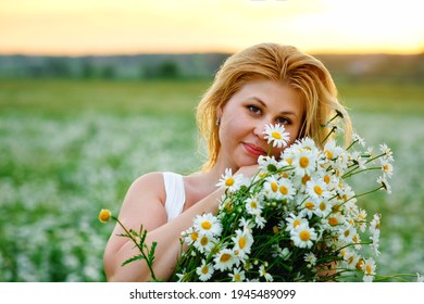 A beautiful plus-size girl with white hair in a summer dress poses outdoors with a bouquet of camomiles. Chubby girl in a meadow with daisies at sunset in the summer.