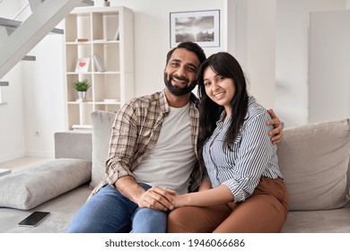 カメラを見て自宅のソファに座って抱き締めて幸せな若いインドのカップルの不動産バイヤー。笑顔の夫と妻の新しい住宅所有者が抱きしめ、自分のアパートの購入を楽しんで、肖像画。