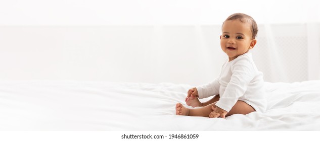 Glad sort lille babypige sidder på sengen og ser på kameraet og smiler i soveværelset derhjemme. Annoncebanner med sødt barn, der poserer indendørs. Panorama med kopiplads til din tekst