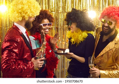 Group of happy friends in retro outfits, feather boas and Afro hair wigs with cake and champagne congratulating young black woman at 70s disco music nightclub fancy dress fashion themed birthday party