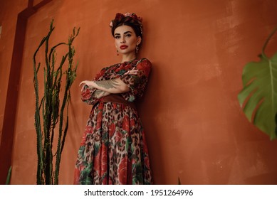 Retrato de una hermosa mujer de cabello oscuro con peinado trenzado de corona y banda de cabeza floral con vestido de gasa elegante con cinturón vasco de pie en la pared étnica naranja entre cactus. estilo mexicano