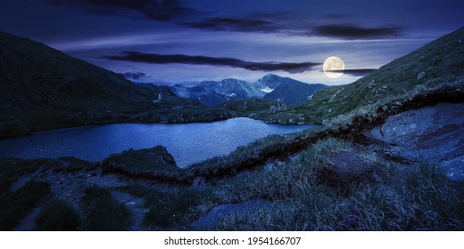 summer landscape with lake on high altitude at night. beautiful scenery of fagaras mountain ridge in summer. open view in to the distant peak beneath a fluffy clouds in full moon light
