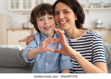 Mom and son making heart hand gesture together happy smiling look in camera at home sitting on sofa. Cute preschool boy and mother take funny family picture photo. Love and happy parenthood concept