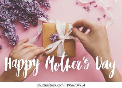 Gelukkige moederdag wenskaart. Happy mother's day tekst en handen inpakken geschenkdoos met lint en lila bloemen op roze papier. Stijlvolle bloemengroeten. Handgeschreven letters. Moederdag