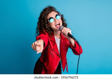 Foto de una dama loca y despreocupada con la boca abierta y cantando, sostenga el dedo directo del micrófono sobre usted, use gafas de sol redondas aisladas en un fondo de color azul