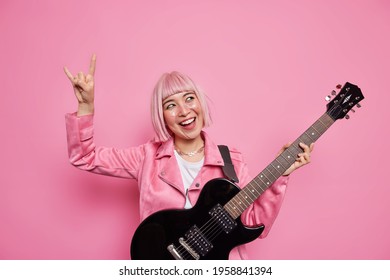 エネルギッシュな幸せなロックスターは、腕を上げたままにし、ピンクの背景にジャケットを着たアコー​​スティックギターを弾き、人気のある曲で自分のアルバムを書くことを幸せにします。陽気なフロントウーマン