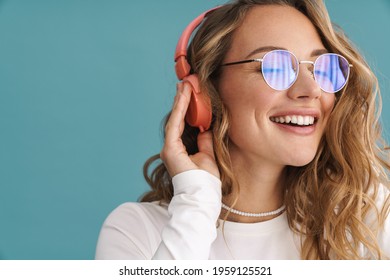 Sonriente joven rubia con gafas escuchando música con auriculares inalámbricos sobre la pared azul