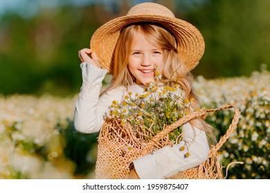 Schattig klein blond meisje in een katoenen jurk en strohoed loopt in een veld met madeliefjes en verzamelt ze in de mand