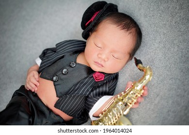 Close up of musician Newborn playing saxophone wearing black suite, black vest and tuxedo hat decorated with red rose. The baby sleeping on grey background.