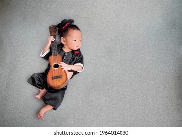 Top van schattige pasgeboren baby's die suites dragen met smokinghoed en rode roos op pak. het kind dat ukulele-gitaar speelt liggend op grijze achtergrondgeluid.
