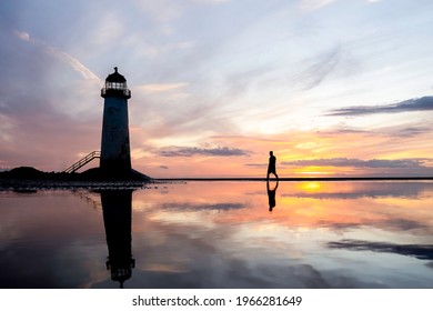水のプールに立っている灯台は、海の水に映る夕焼けの反射を見事に表現しています。ウェールズ北部の海岸の砂のビーチを探索する一人の孤独な男は、静かな水のオレンジ色の輝き日の出ゴールデンアワーブルーアワー.
