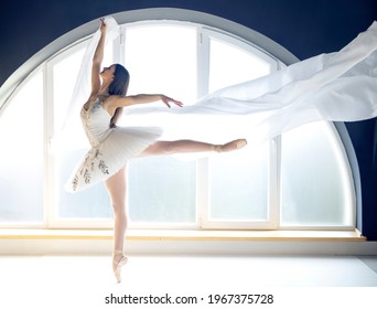 白いチュチュの衣装に身を包んだ若いやる気のあるバレリーナは、自然の周囲の昼光の雰囲気の中で、大きな丸い形の窓の背景の前のバレエスタジオでバレエの練習をします