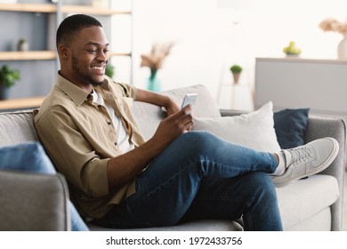 Excelente aplicación móvil. Hombre afroamericano feliz usando el teléfono, enviando mensajes de texto y navegando por Internet sentado en el sofá de su casa, vista lateral. Hombre negro que se conecta en las redes sociales usando una aplicación en un teléfono inteligente